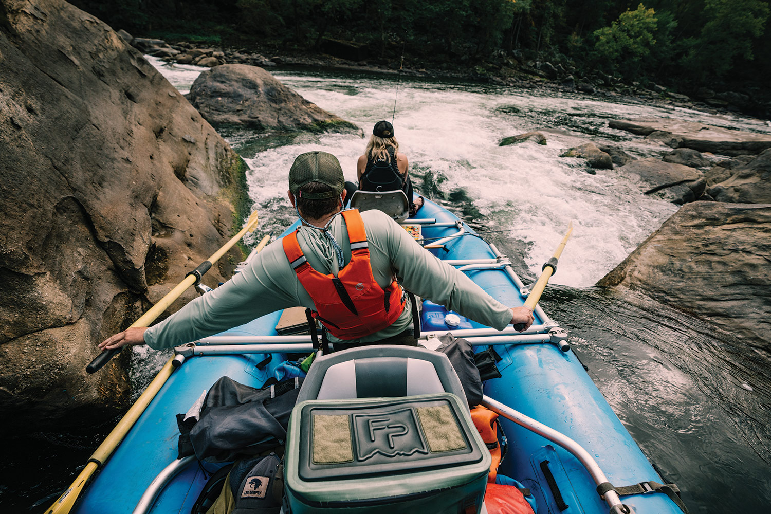 Rafter faces forward to guide raft downriver through rocky rapids; angler sits in front of boat.