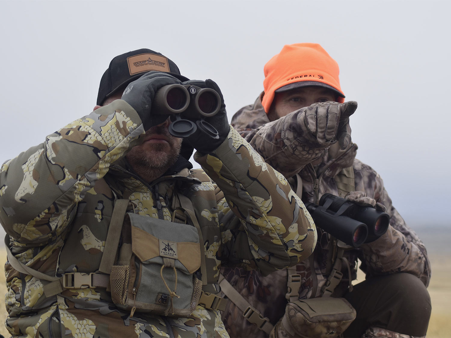 Two hunters scout a plain. One uses binoculars while the other points in a direction to scout.