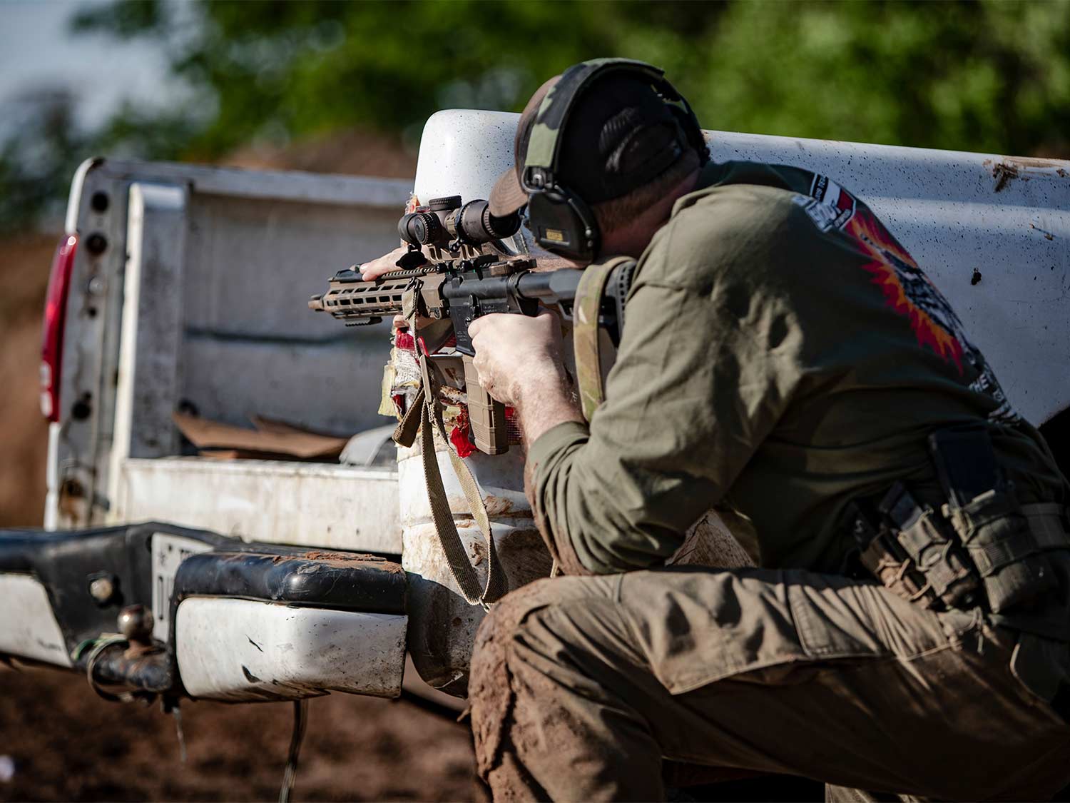 A man aims a rifle while crouched behind the bed of a truck.