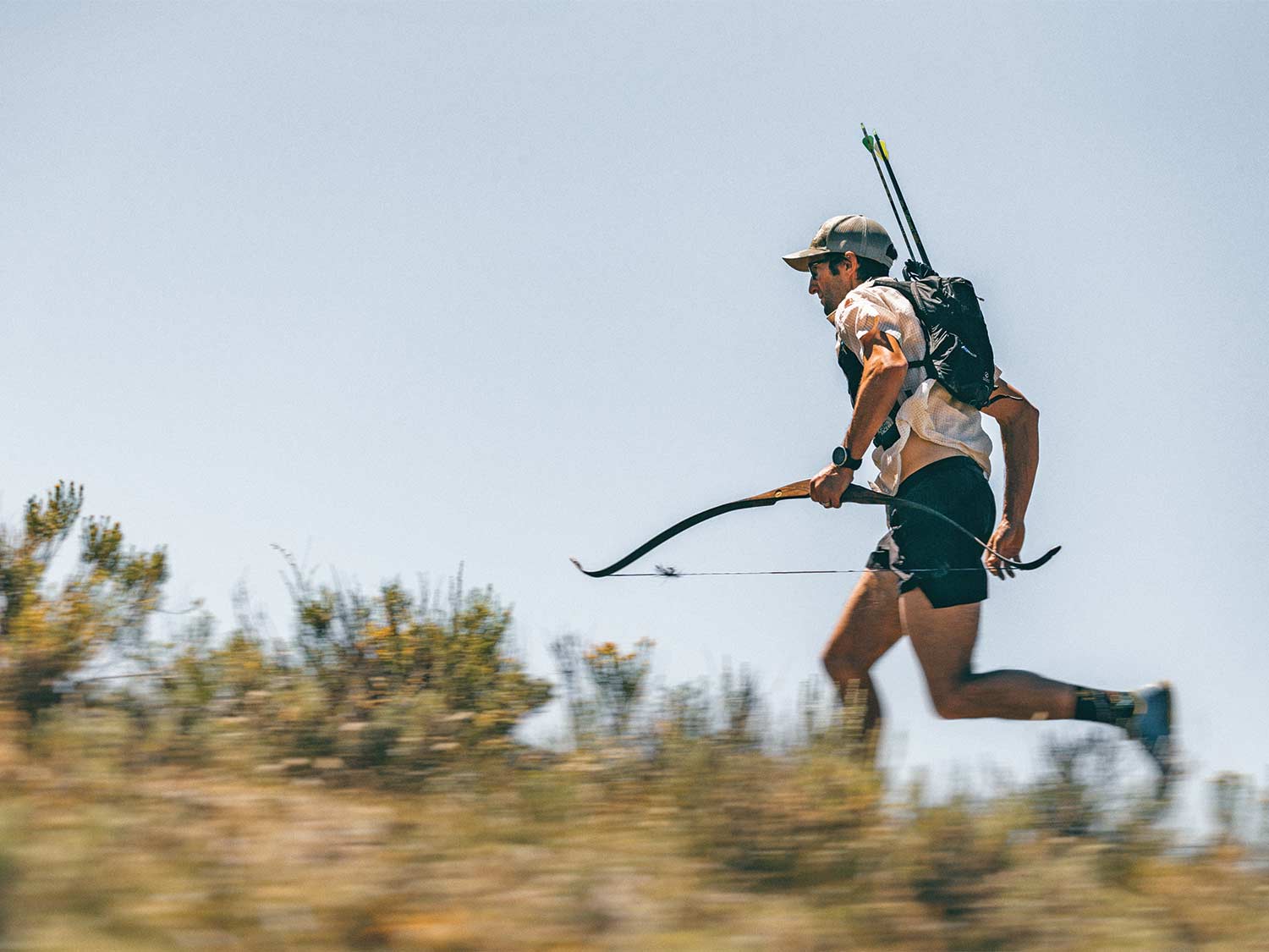 A hunter holds a traditional bow and runs through an open field.