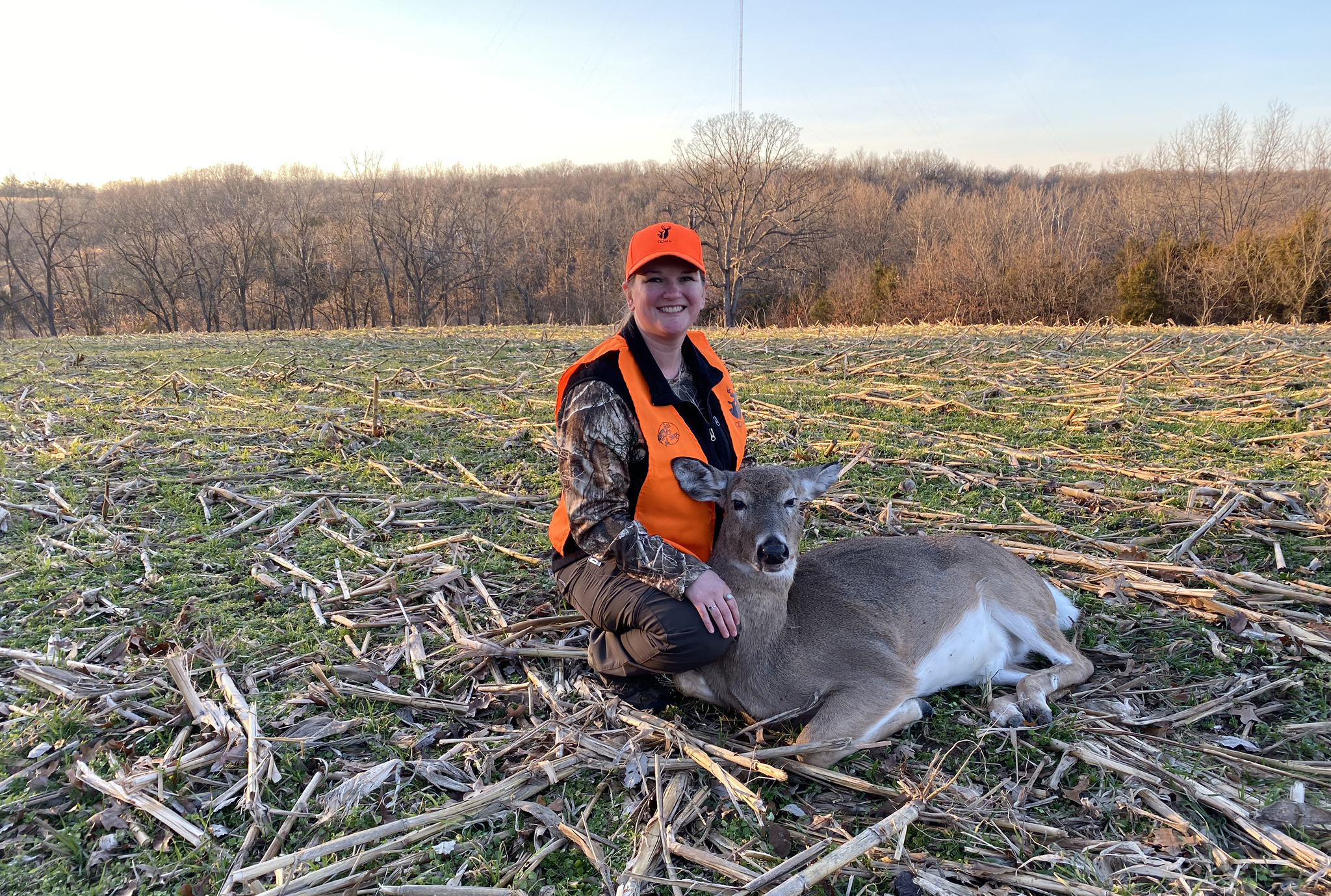 A female hunter in blaze orange kneels beside a doe in a cut cornfield in early winter.