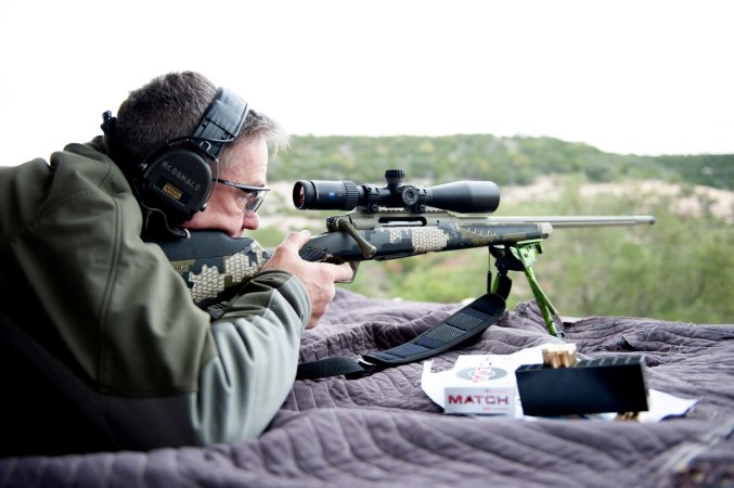 A man with glasses and ear protection lying prone behind a camo rifle on a shooting blanket in rugged country.