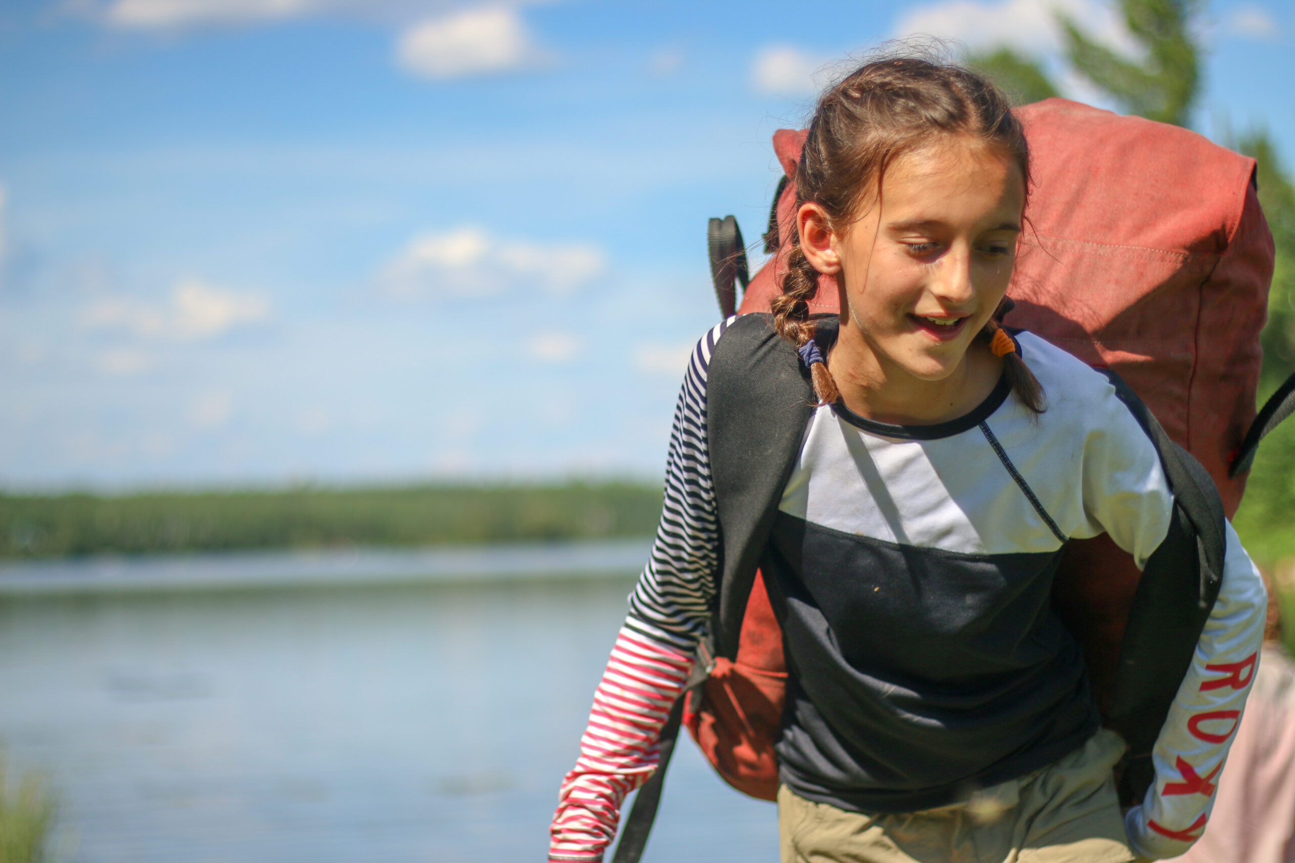 A girl shoulders her pack on the shore of a northern lake.