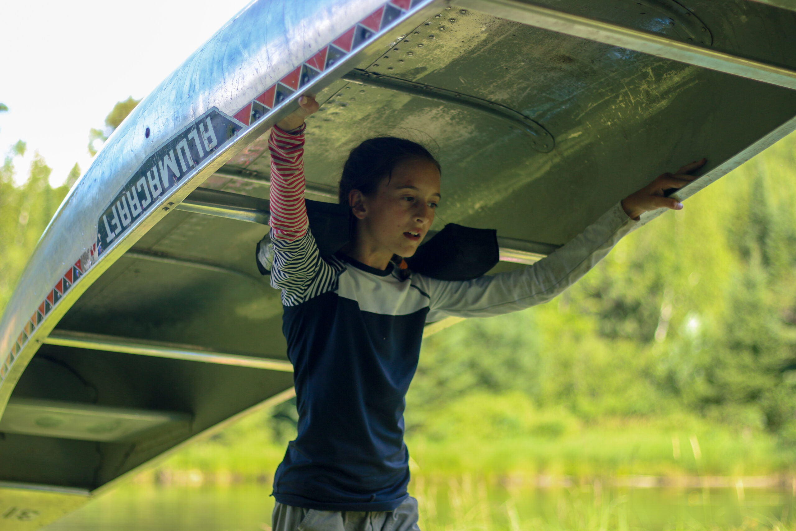 A girl carries an aluminum canoe on her shoulders during a portage.