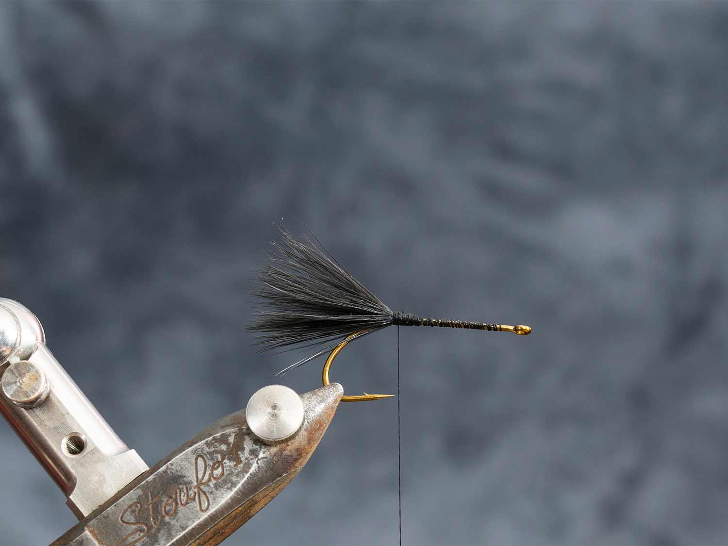 A fly lure hook clamped in a vice while someone adds more attachments and adjustments to build a custom lure.