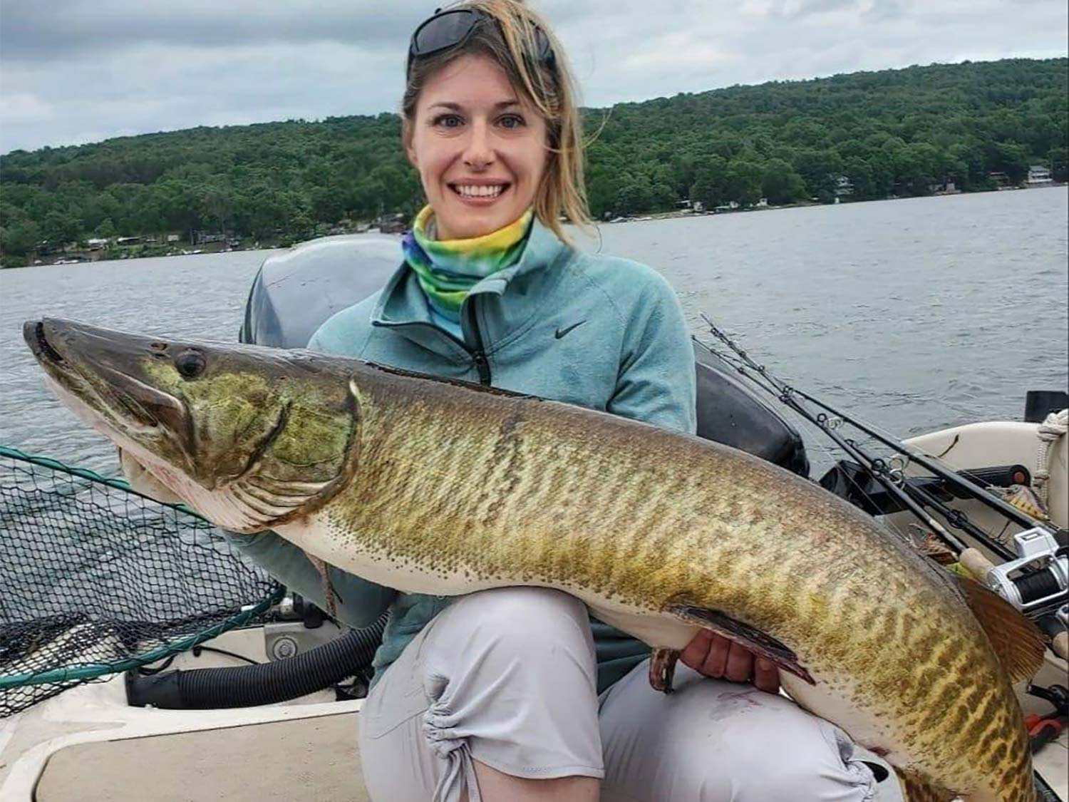 A woman smiles and poses while holding a large muskie fish.