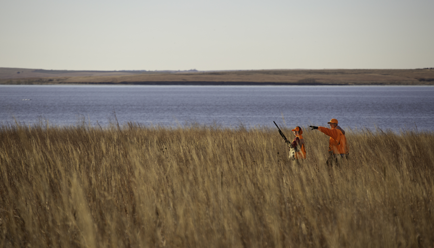 An older upland hunter in blaze orange points out something to a new hunter.