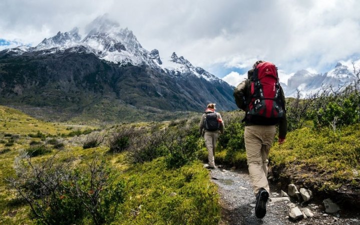 Two people with camping backpacks walking through a green field among the mountains.