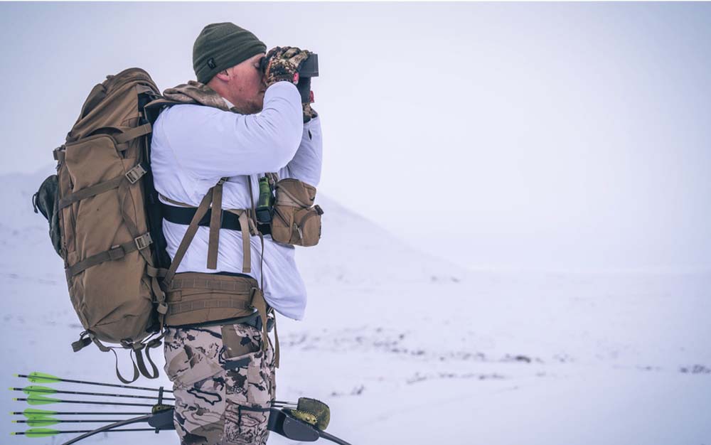 A man in the snow wearing a backpack looking through his binoculars