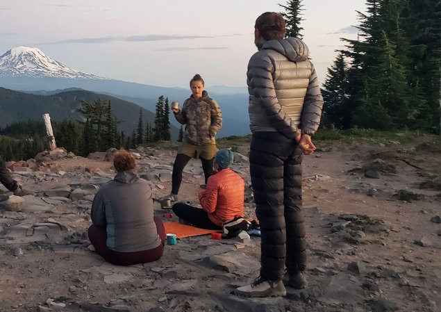 man standing in down pants in front of a crowd facing a mountain at sunset