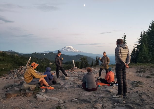 group of people sitting around on a rocky field with a volcano in the background at sunset