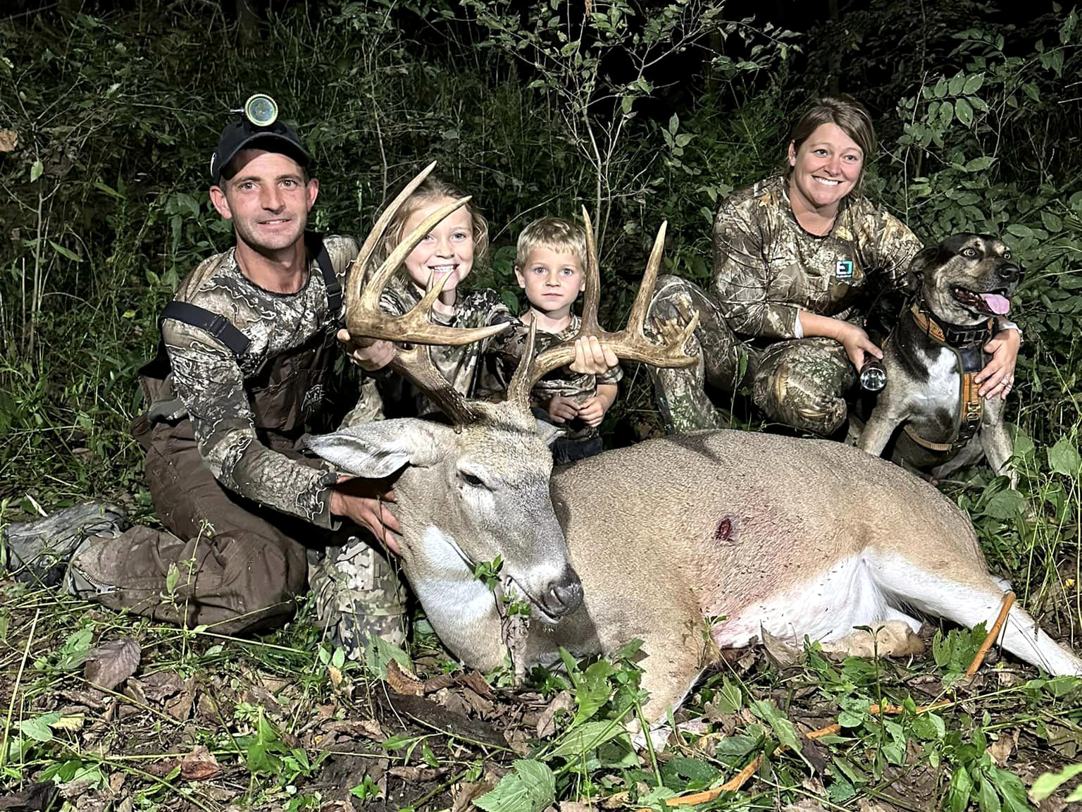 A family and their tracking dog sit behind a big buck.