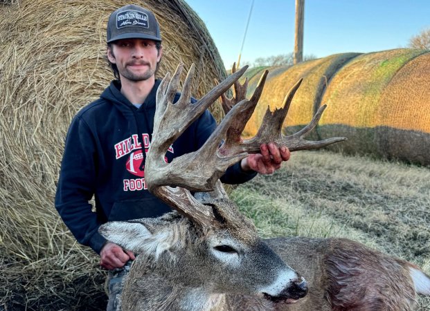 A hunter sits with a nontypical buck in front of a hay bale.