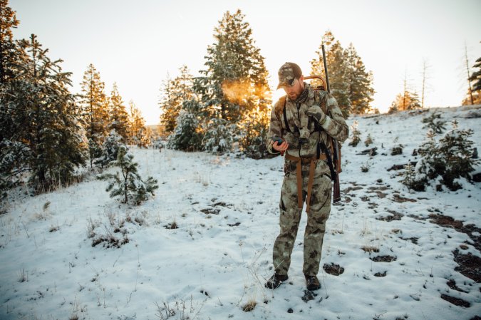 A hunter in the snow looks at his phone.