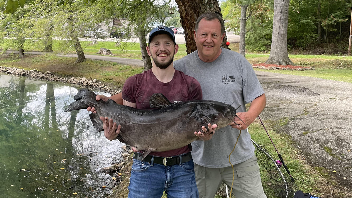 Two men smile while one holds a giant catfish.