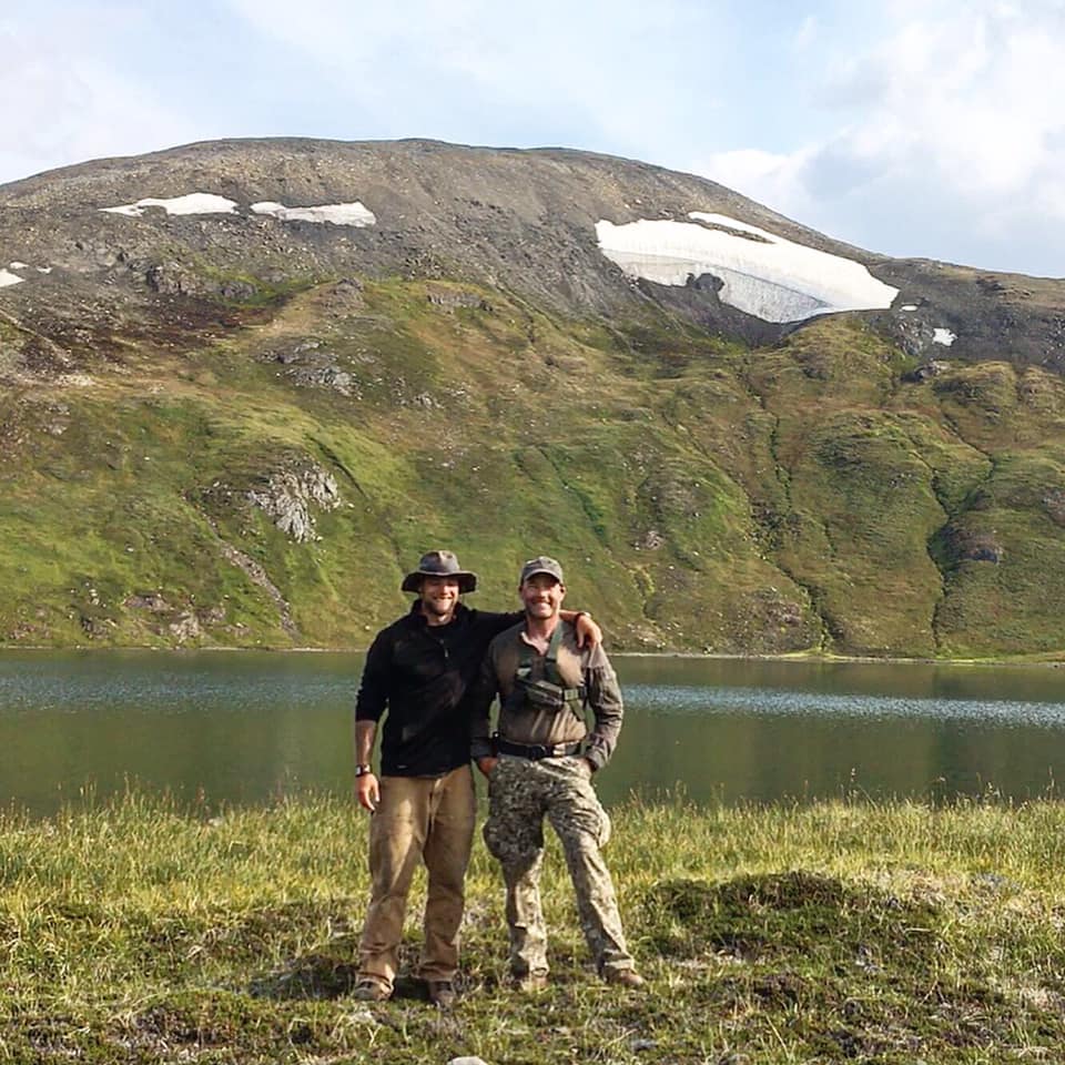 Two men pose in front of a stream in the mountains