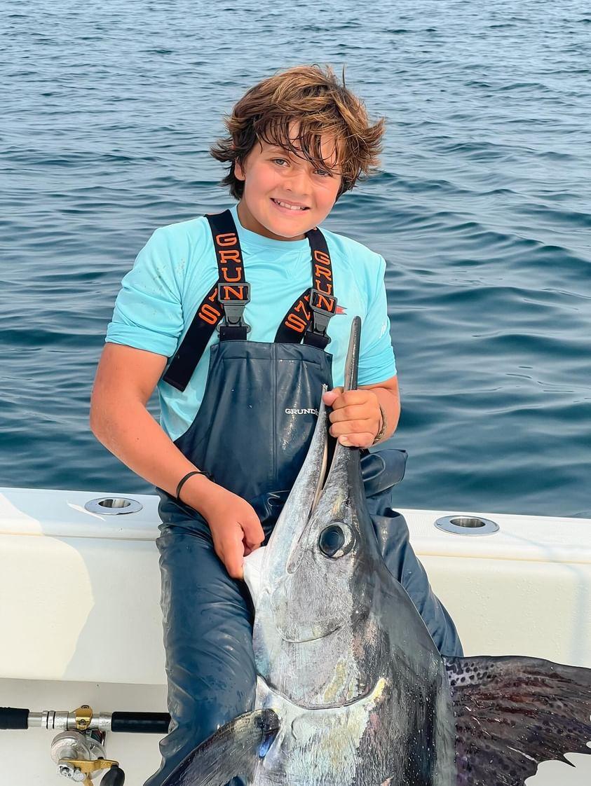 A kid in Grundens waders holds up a white marlin.