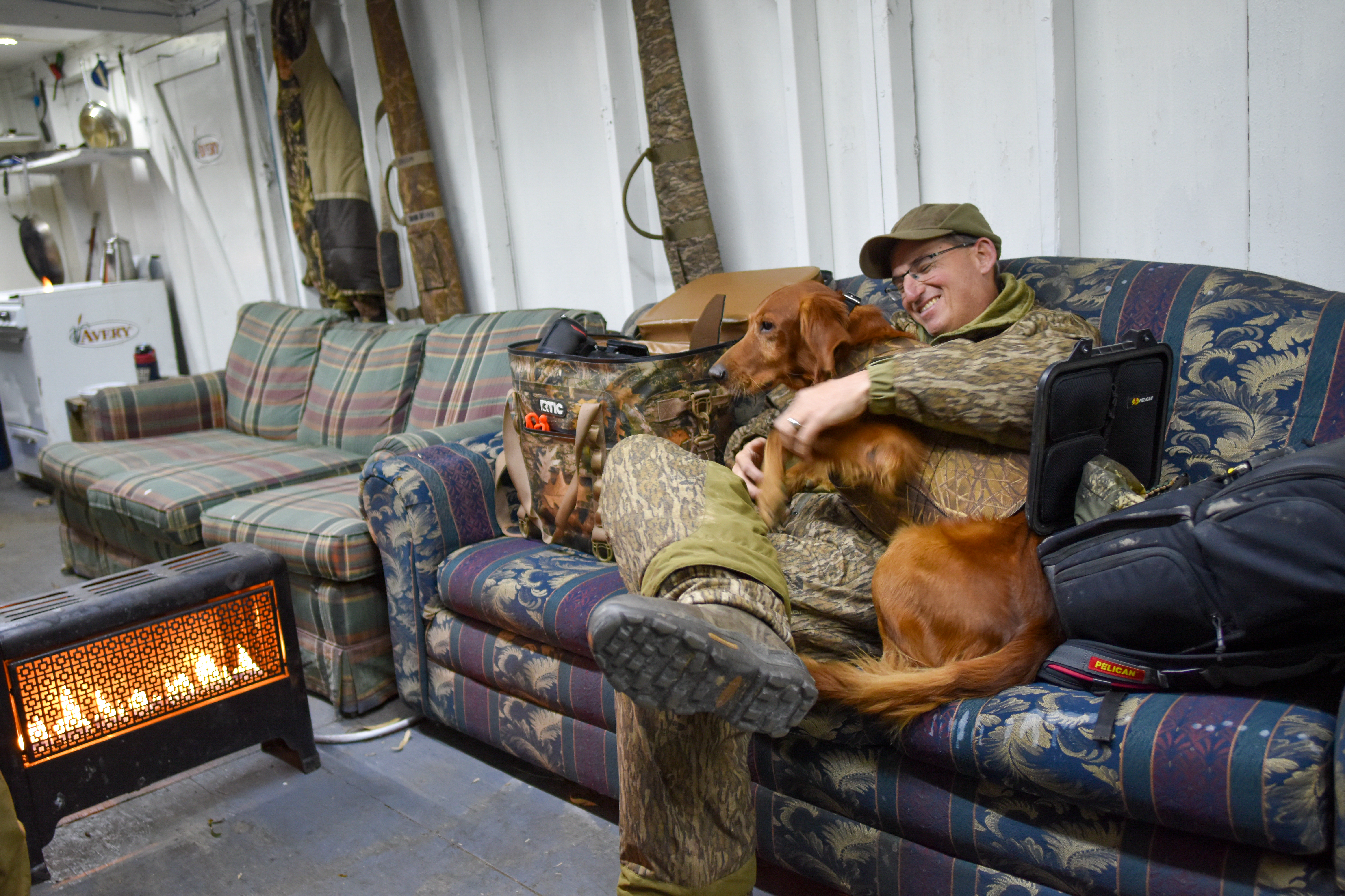 A hunter sits on a couch with a dog in a fancy duck blind.