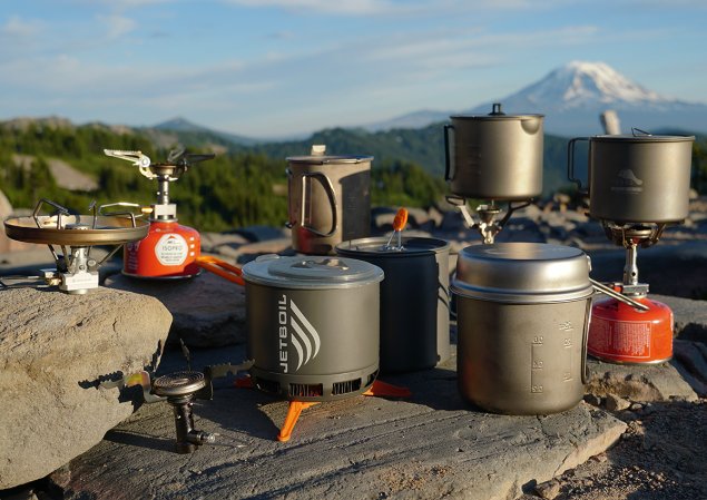 a variety of backpacking stoves and backpacking pots set up against a mountain backdrop