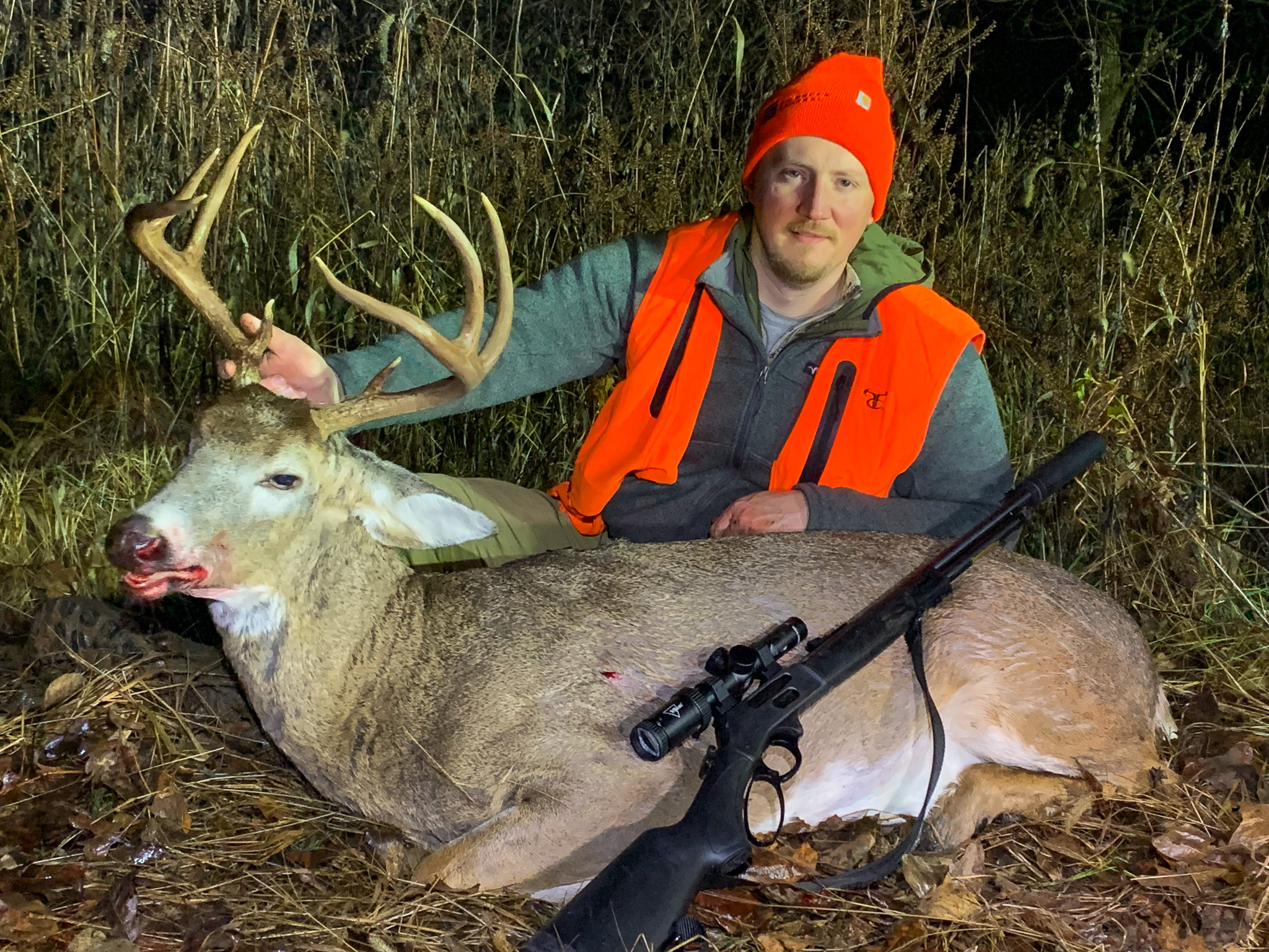 Hunter Tyler Freel in blaze orange sits behind a whitetail buck with a suppressed lever action rifle resting on it.