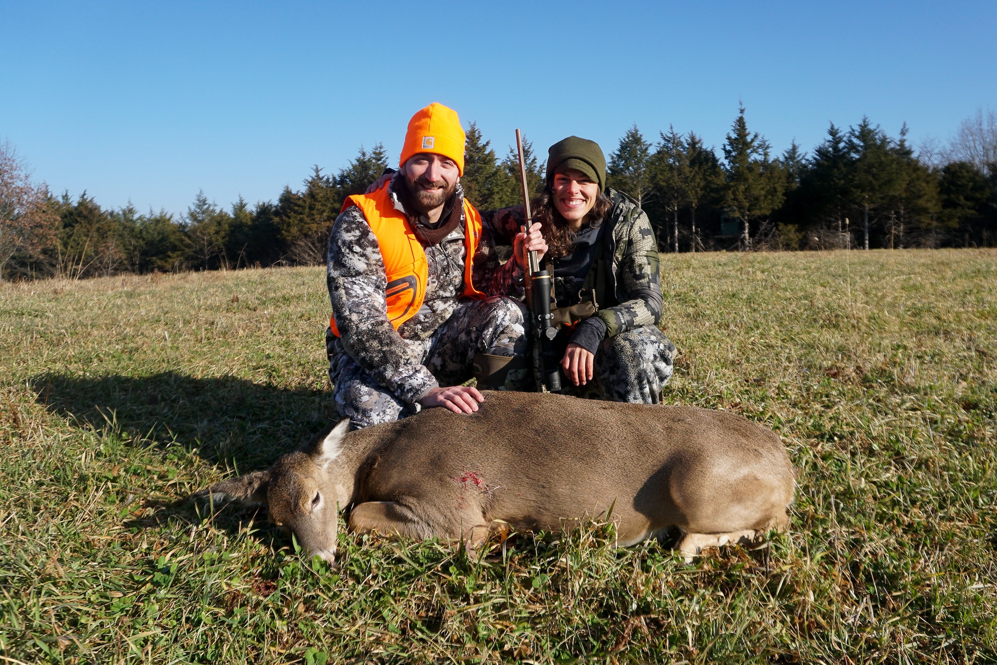 Two hunters, one in blaze orange, kneel behind a dead doe.