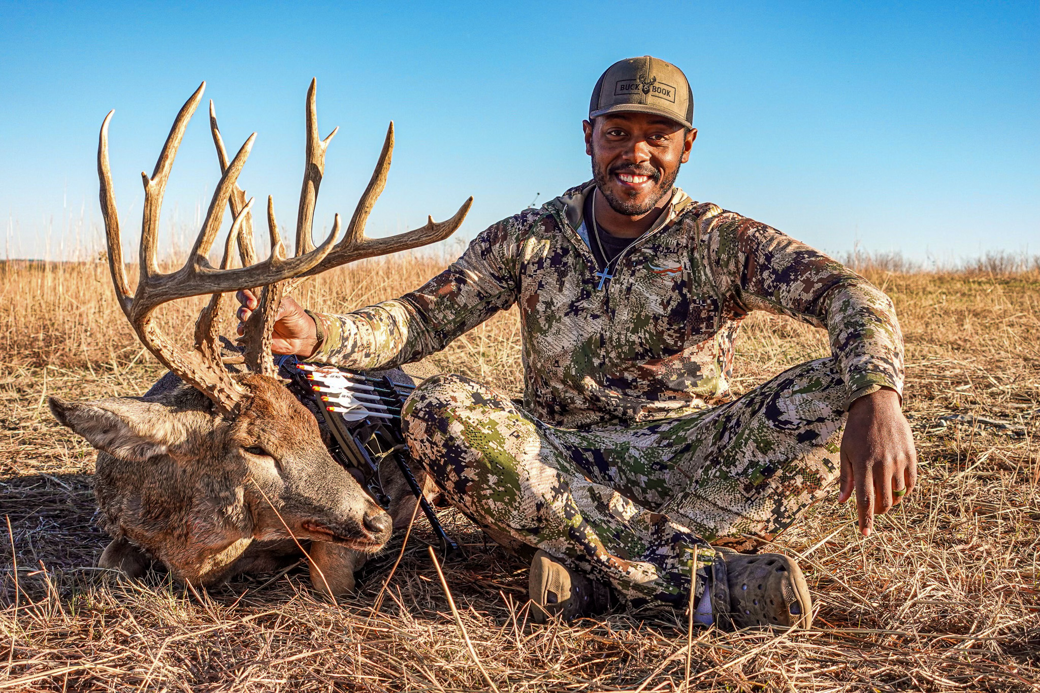 A bowhunter sitting crosslegged in a field beside a big buck