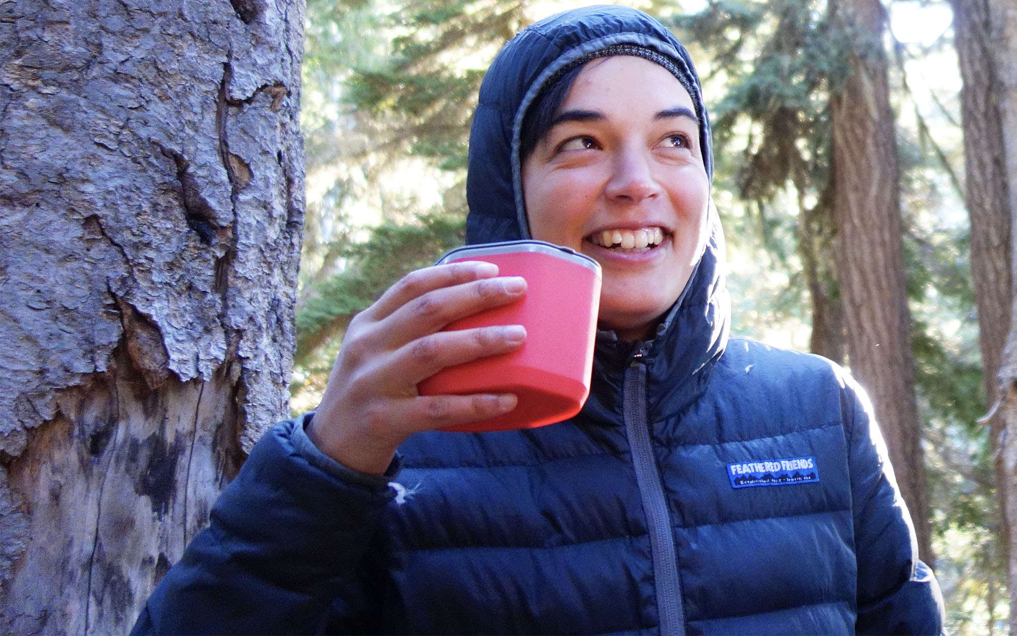 women wearing a hooded puffer jacket in the woods drinking out of a red mug