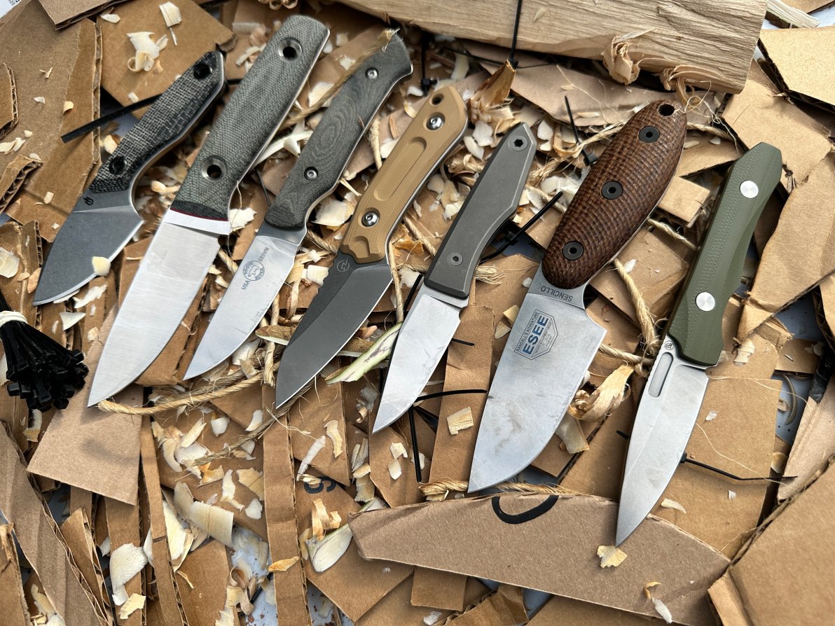 A bunch of fixed blade edc knives lying on sliced cardboard.