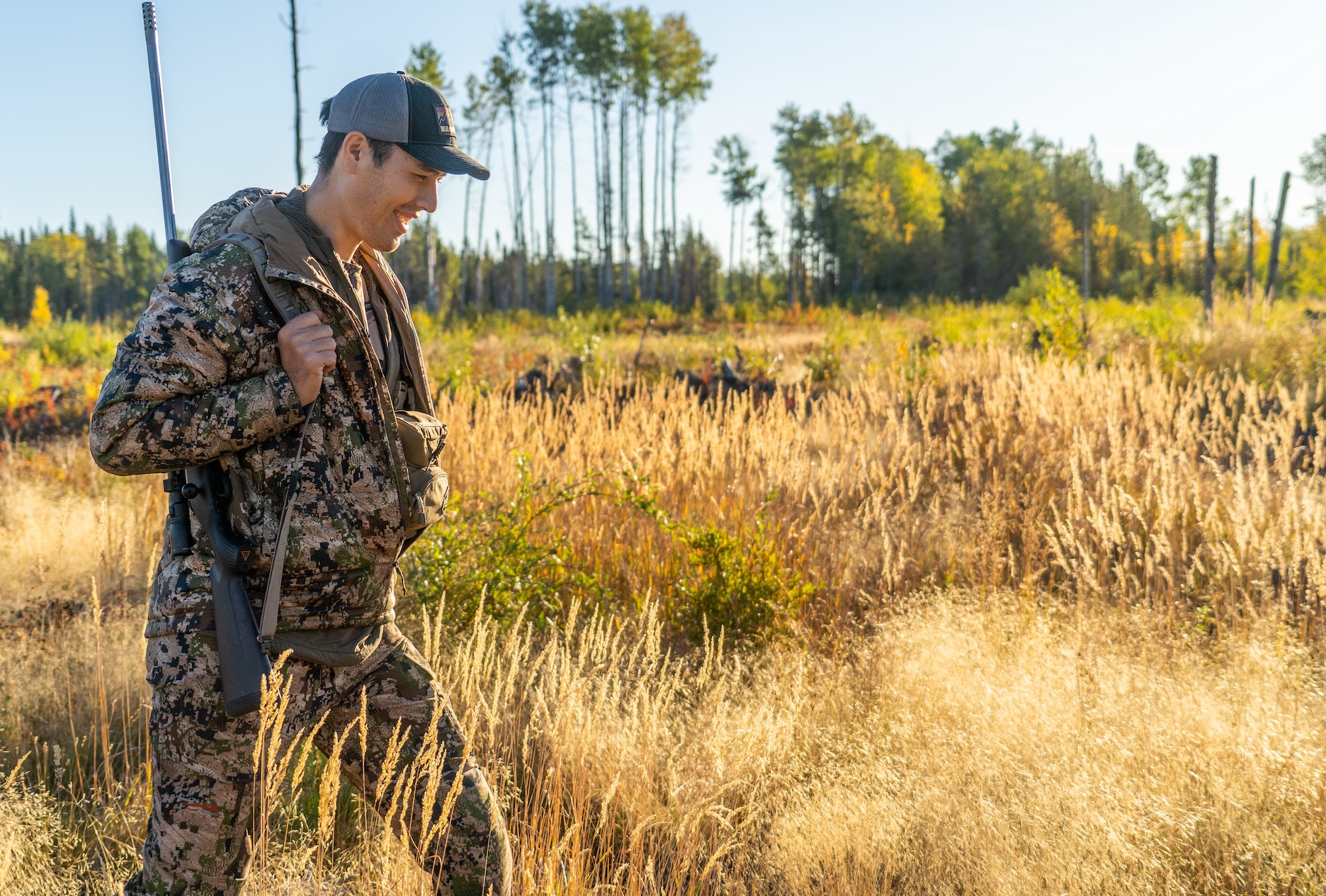 A man dress in camo carrying a rifle walks through a field of tall grass