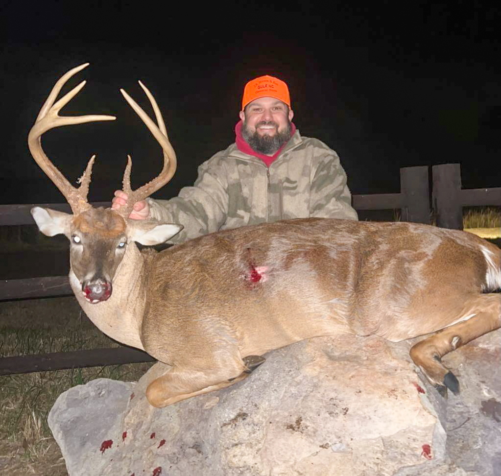 A hunter stands behind a big buck on a rock.