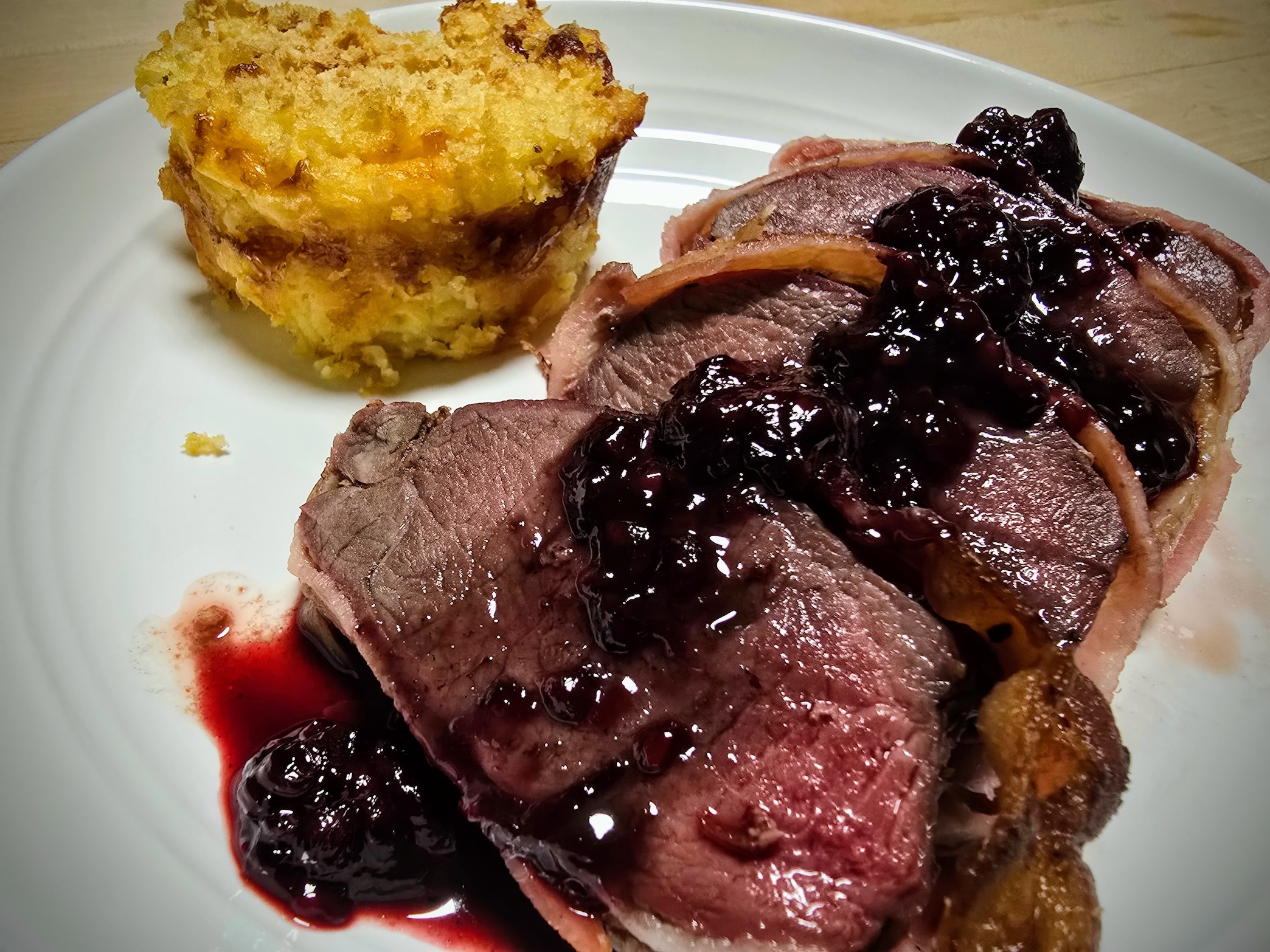 Sliced venison backstrap wrapped in bacon and topped with blackberry sauce sits on a white plate. 