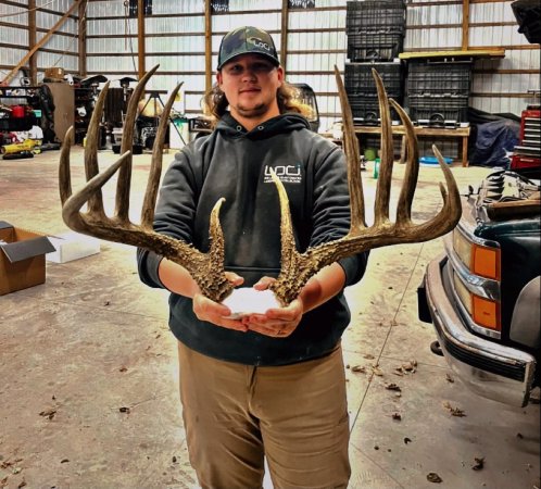 A young man wearing a ball cap stands in a garage holding a large set of whitetail deer antlers.