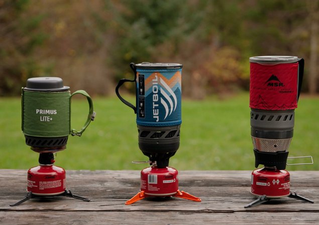 Primus Lite Plus, JetBoil Flash, and MSR Windburner backpacking stoves sitting on a picnic table with a grassy field in the background