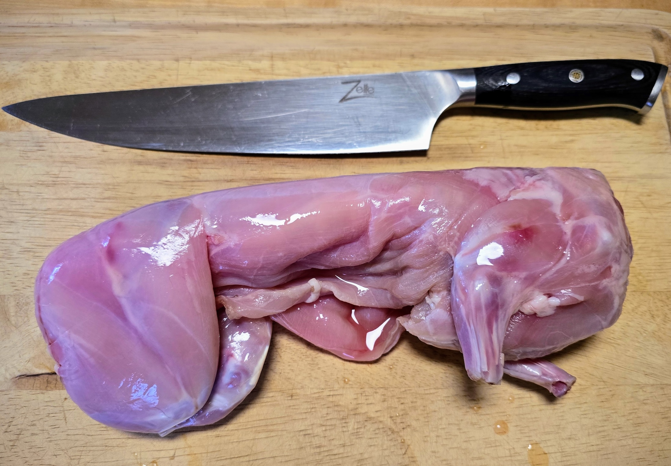 A skinned rabbit sits on a wooden cutting board next to a black-handled chef's knife.