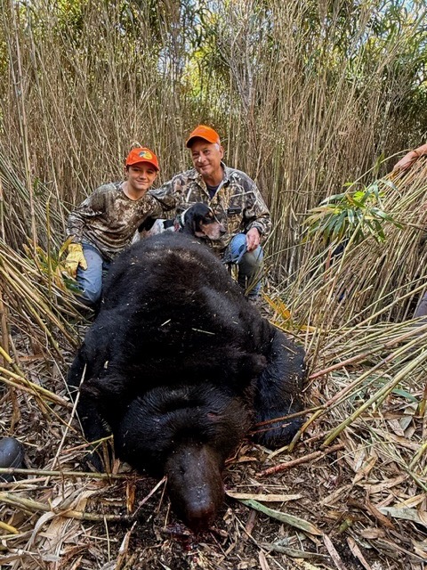 A young boy and an older man in blaze orange hats pose with a hound dog and a large black bear in high reeds.