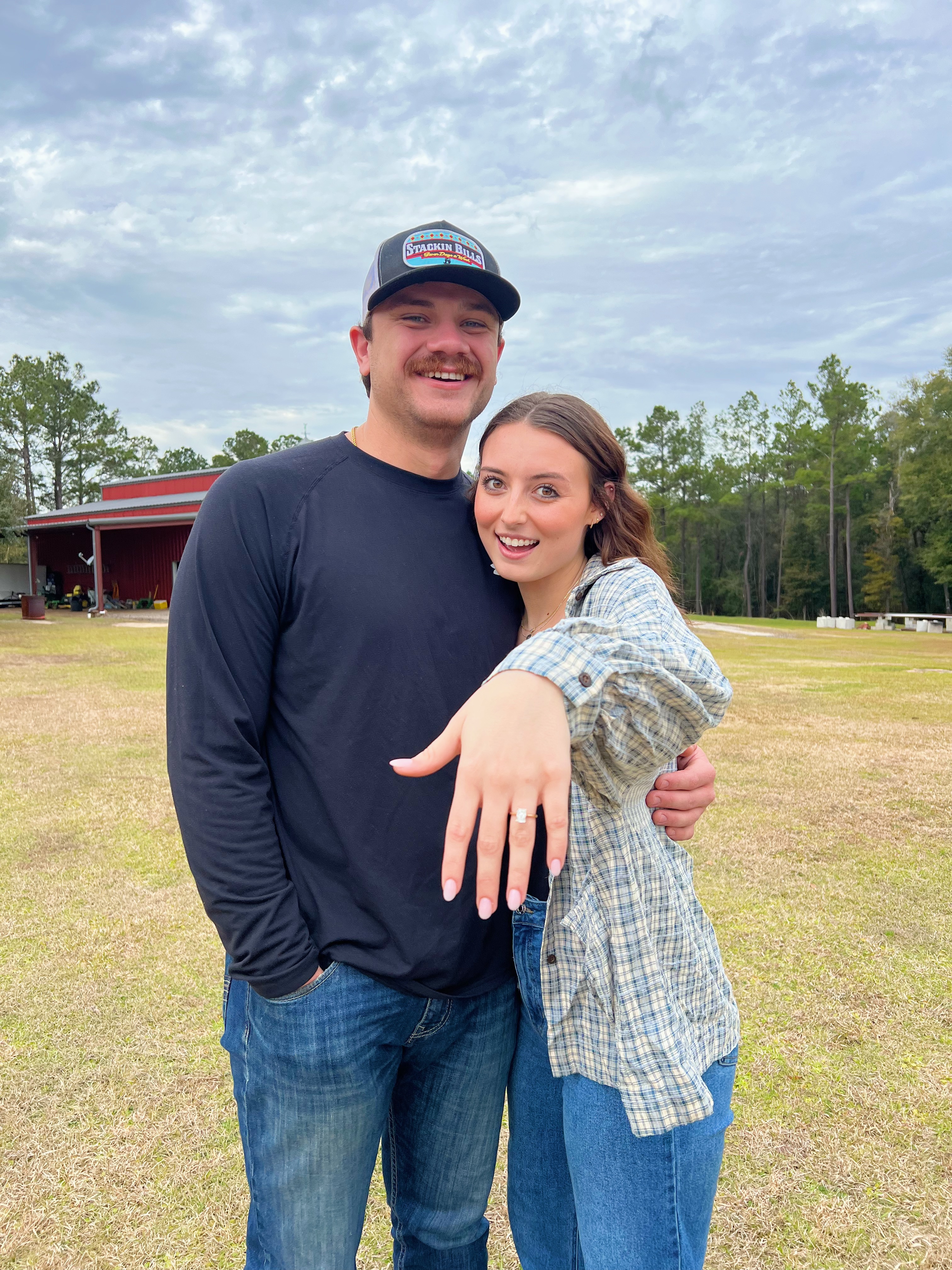 A man and woman stand side by side smiling as the woman shows off an engagement ring.