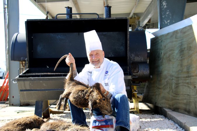 A chef poses with a giant nutria in front of a large barbecue grill.